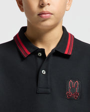 A person wearing a Psycho Bunny KIDS PIERCE PIQUE POLO in black, showcasing red detailing on the collar and an embroidered bunny skull logo on the chest. The photo focuses on the upper chest and lower face, featuring a neutral expression.