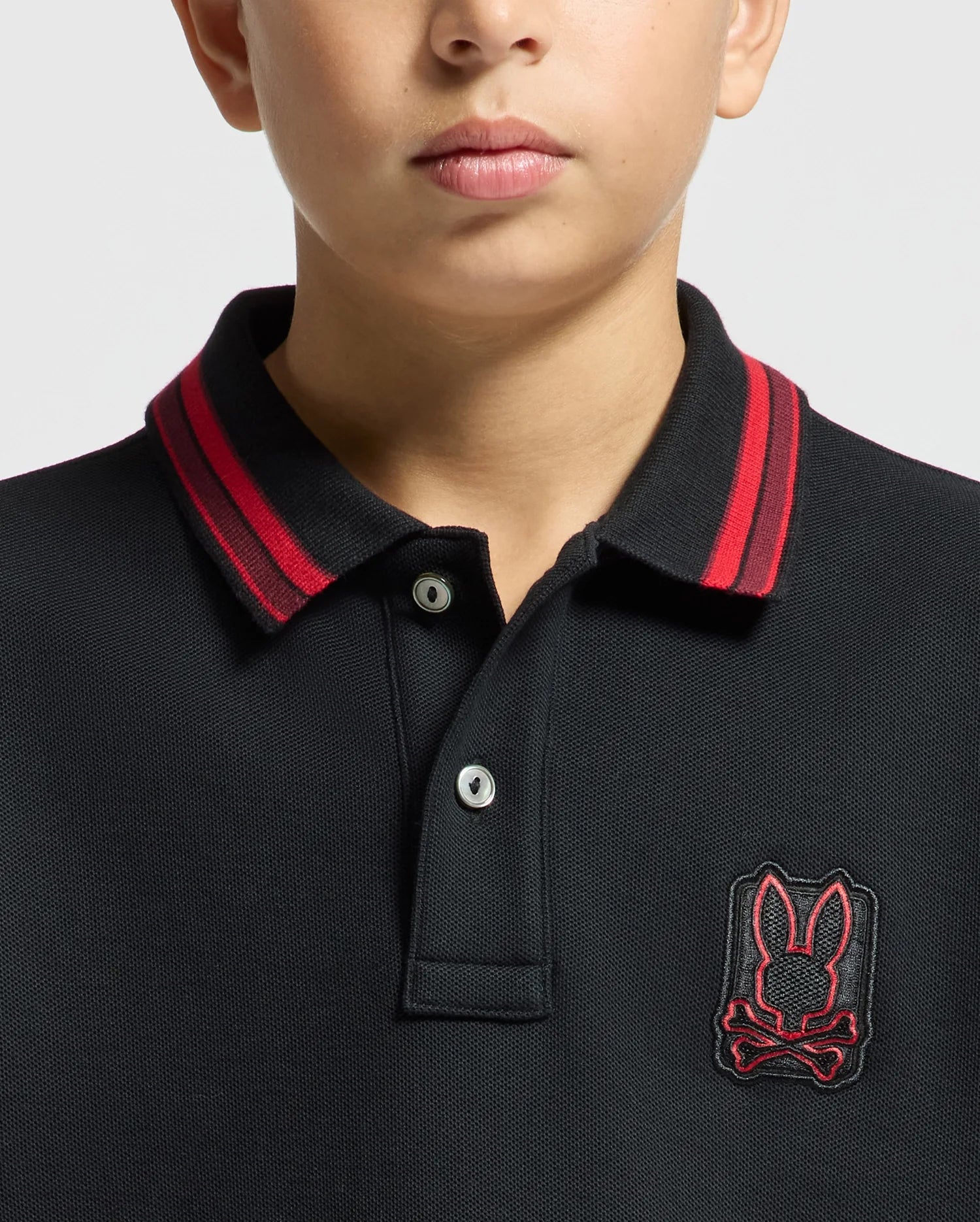 A person wearing a Psycho Bunny KIDS PIERCE PIQUE POLO in black, showcasing red detailing on the collar and an embroidered bunny skull logo on the chest. The photo focuses on the upper chest and lower face, featuring a neutral expression.