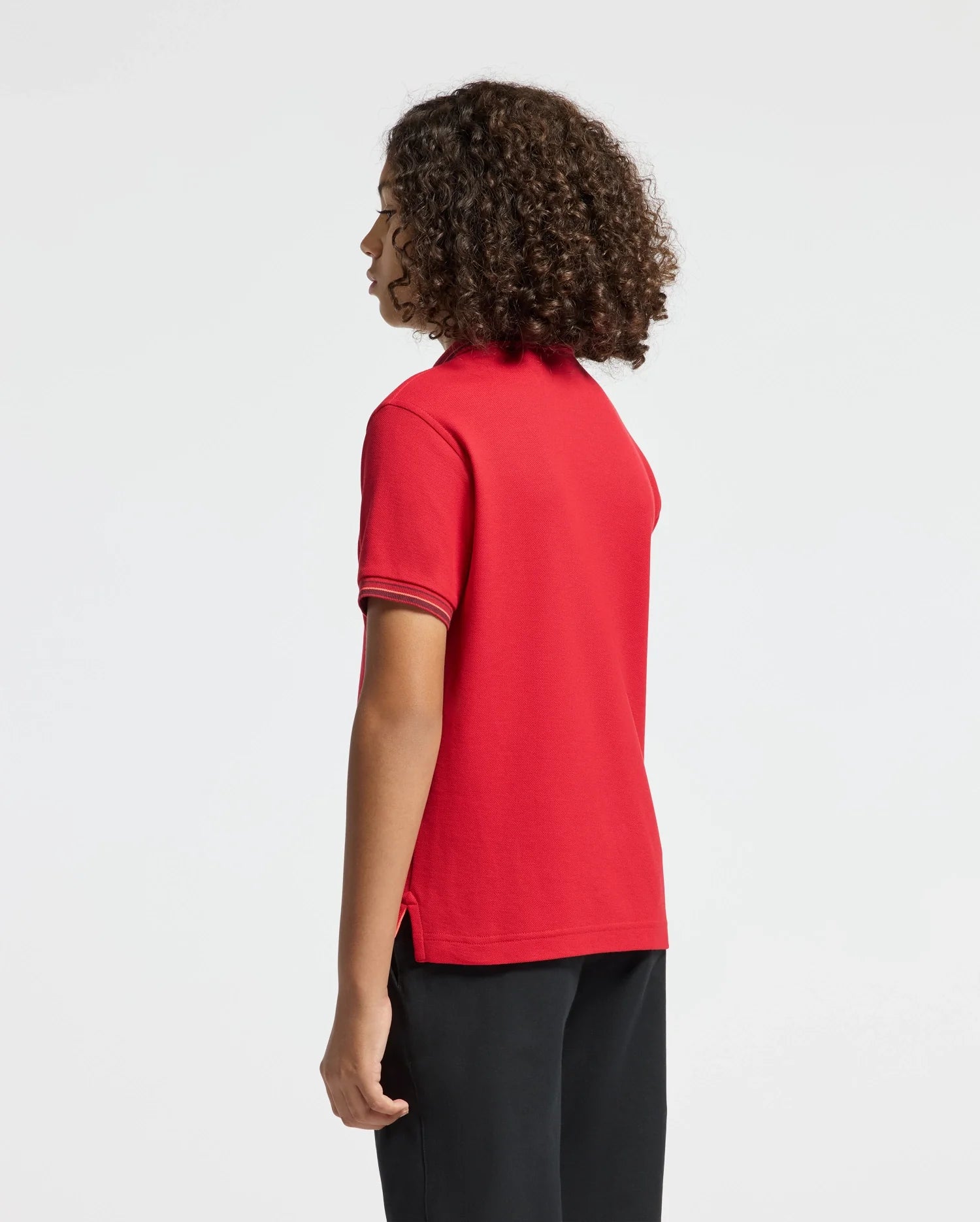 A person with curly hair is dressed in a red KIDS PIERCE PIQUE POLO - B0K201E200 by Psycho Bunny, showcasing an embroidered chest Bunny. They pair it with black pants and stand gracefully, gazing to the side against a plain background.