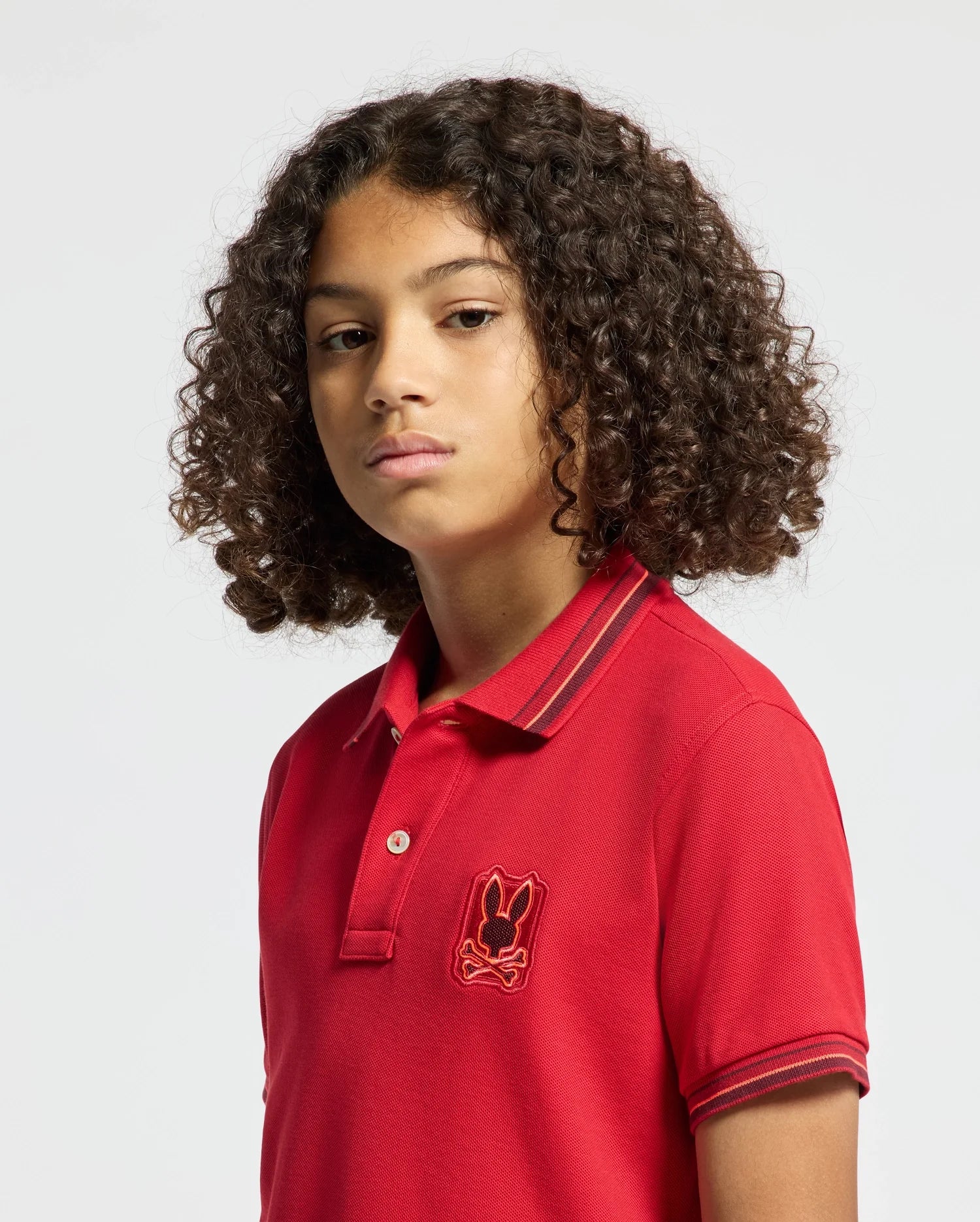 A young person with curly hair stands against a plain background, wearing the KIDS PIERCE PIQUE POLO - B0K201E200 by Psycho Bunny, which showcases an embroidered chest Bunny. They have a neutral expression.