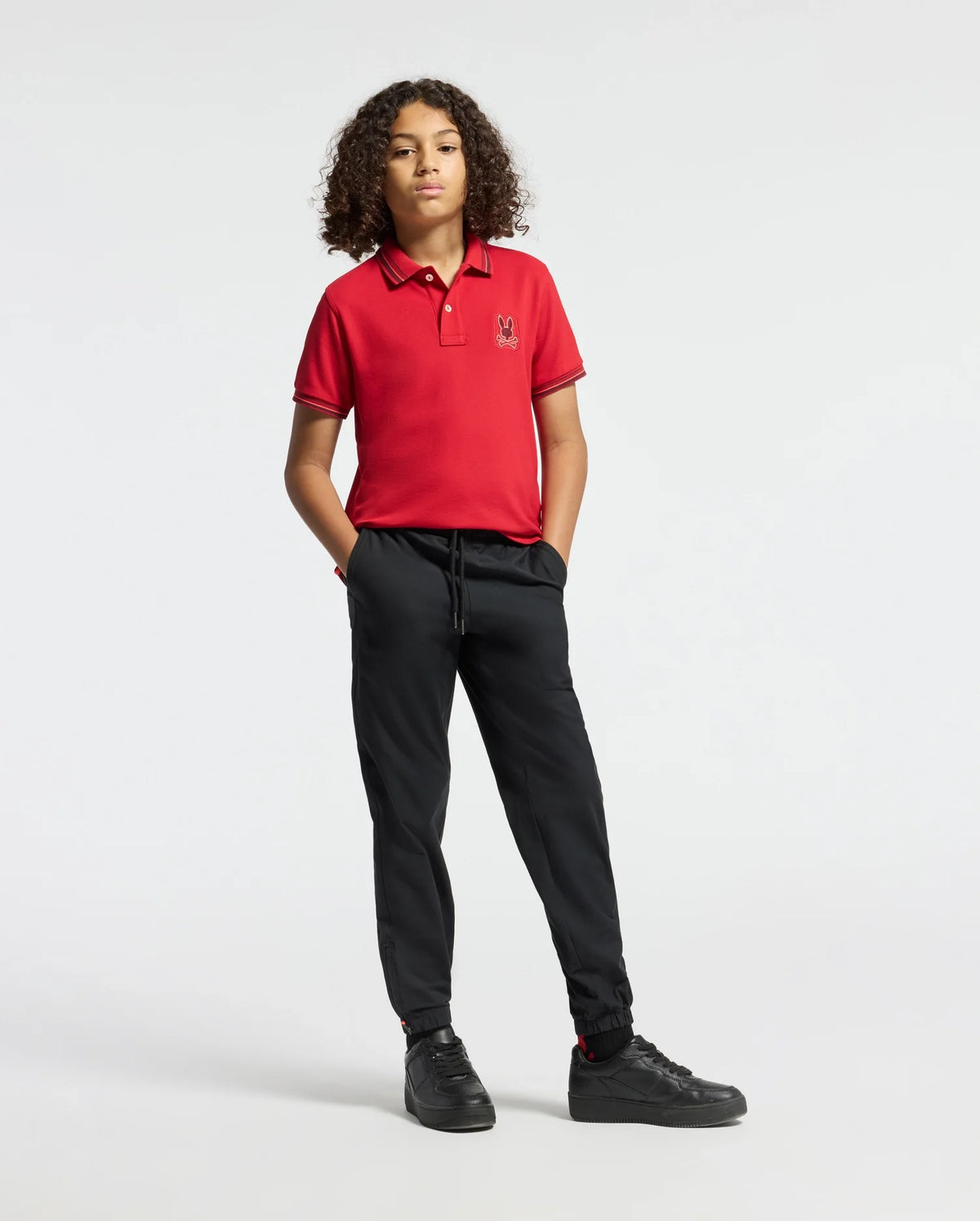 A young individual with curly hair exudes confidence, dressed in the Psycho Bunny KIDS PIERCE PIQUE POLO - B0K201E200, a fashionable red polo shirt paired with black pants and hands in pockets. The embroidered chest Bunny design introduces a whimsical element, while black shoes complement the ensemble against a plain white backdrop.