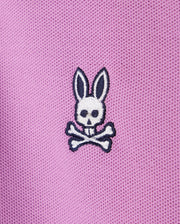 A close-up of pink Psycho Bunny KIDS SOUTHPORT PIQUE POLO SHIRT - B0K263B200 fabric with a small embroidered white bunny skull and crossbones design. The bunny has long ears, black eyes, and black detail on the ears and skull. The fabric, reminiscent of a kids polo, has a textured, mesh-like appearance.