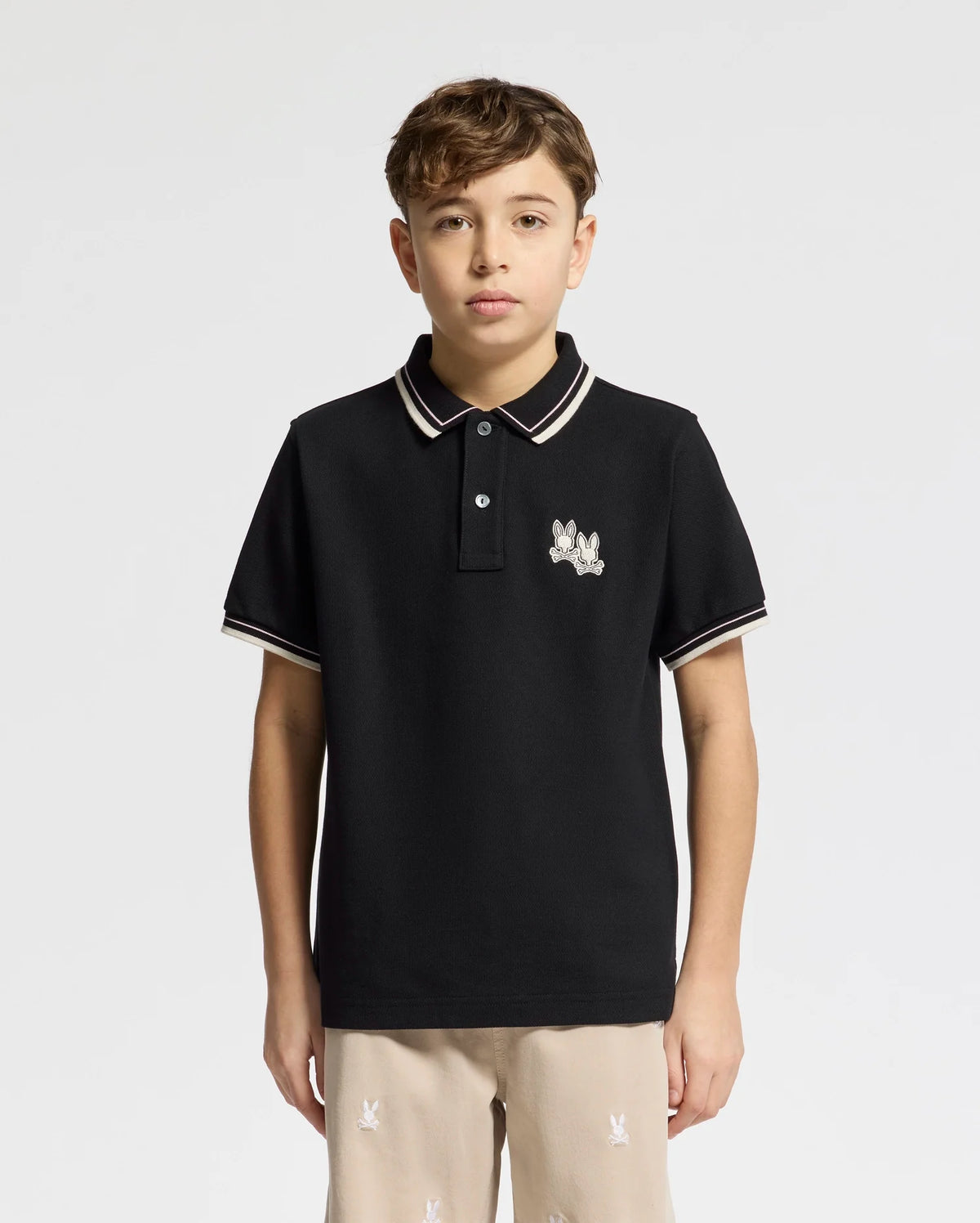 A young person models the black Psycho Bunny KIDS ELLIOT PIQUE POLO (B0K505F200), featuring white trim and an embroidered chest logo, against a plain white background. They have short brown hair and are looking directly at the camera, highlighting the polo's refined details.