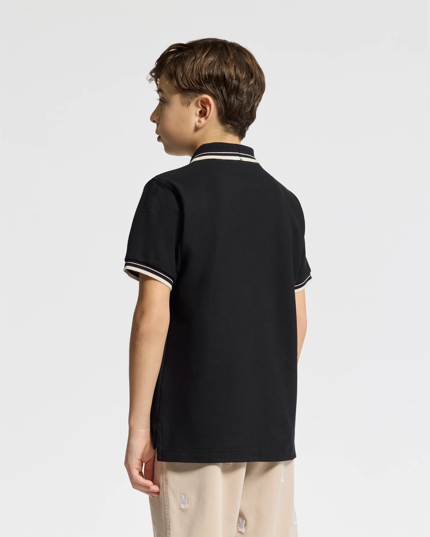 A young boy with short brown hair wears the Psycho Bunny Kids Elliot Pique Polo in black, featuring white trim on the collar and sleeves. This pima cotton polo showcases elevated finishes, complementing his beige pants against a plain white background.