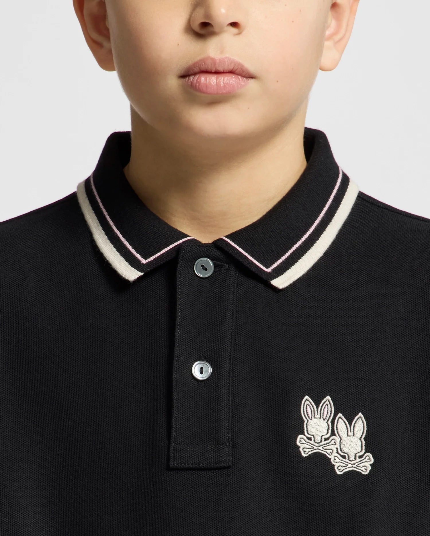 A close-up shows a person wearing the Psycho Bunny KIDS ELLIOT PIQUE POLO (B0K505F200) in black, made from soft Pima cotton with white and pink collar trim, featuring two bunny logos on a plain light gray background.