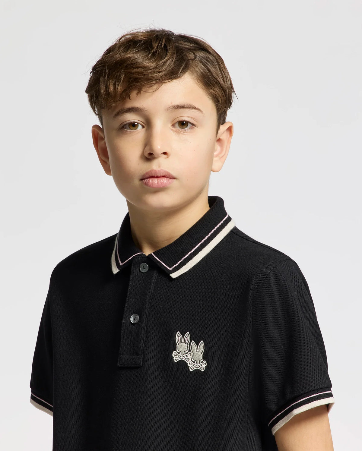A young person with short brown hair wears the black Kids Elliot Pique Polo by Psycho Bunny, featuring embroidered bunny motifs on the left chest and a stylish white and black striped collar. The background is plain light gray.