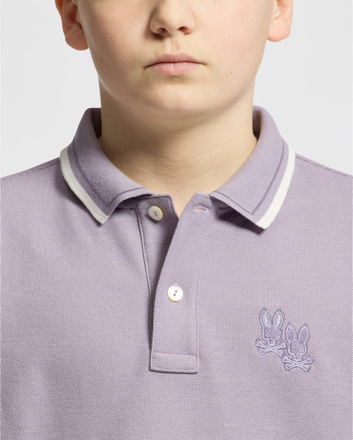 A fashionable individual models a Psycho Bunny KIDS ELLIOT PIQUE POLO in light purple crafted from soft Pima cotton featuring white trim and an embroidered design of two rabbits on the chest against a plain off-white background.