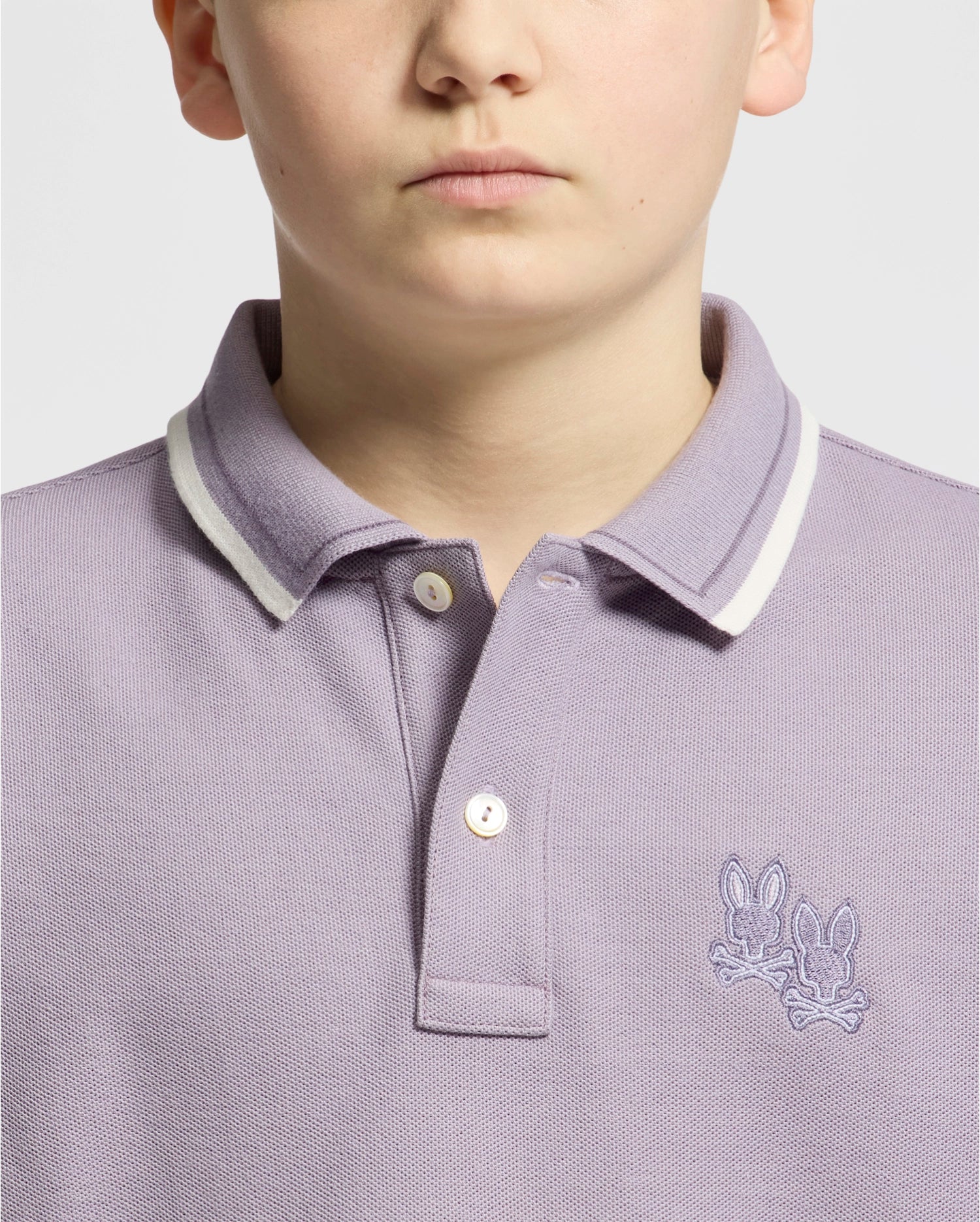 A fashionable individual models a Psycho Bunny KIDS ELLIOT PIQUE POLO in light purple crafted from soft Pima cotton featuring white trim and an embroidered design of two rabbits on the chest against a plain off-white background.