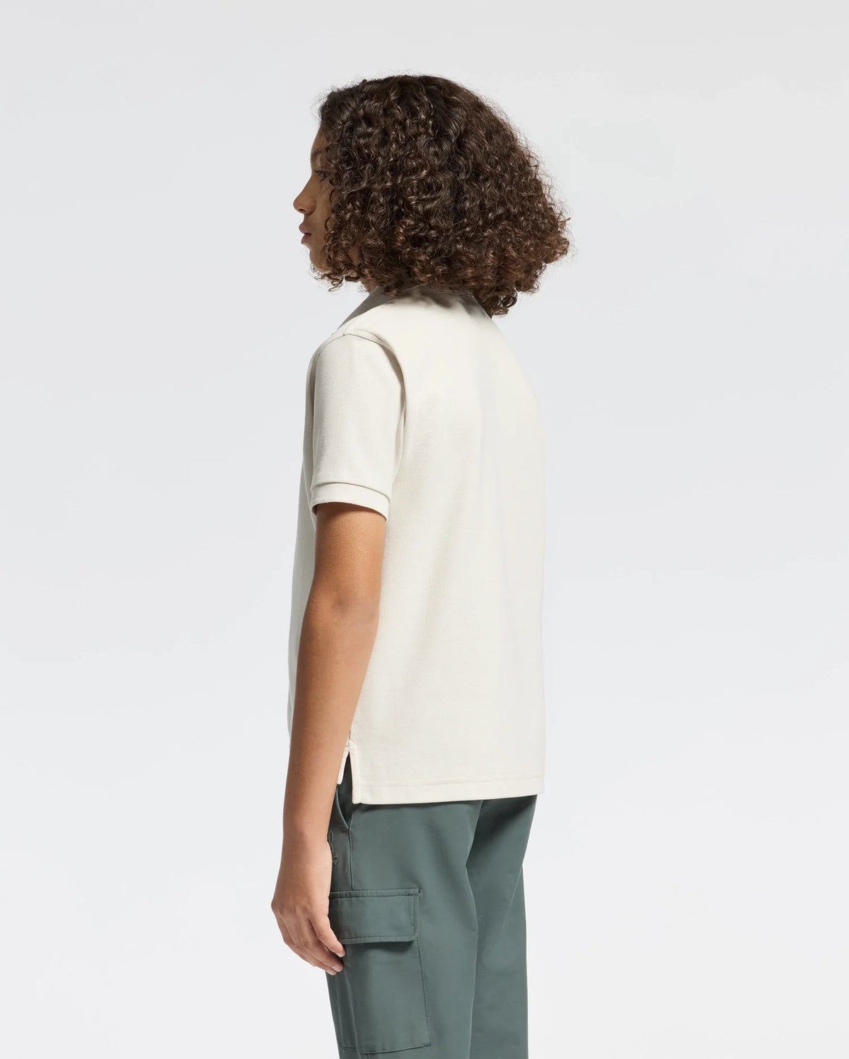 A person with curly hair stands sideways in a Psycho Bunny KIDS DESERT PIQUE POLO - B0K506F200 and dark green cargo pants, exuding timeless style against a plain white background.