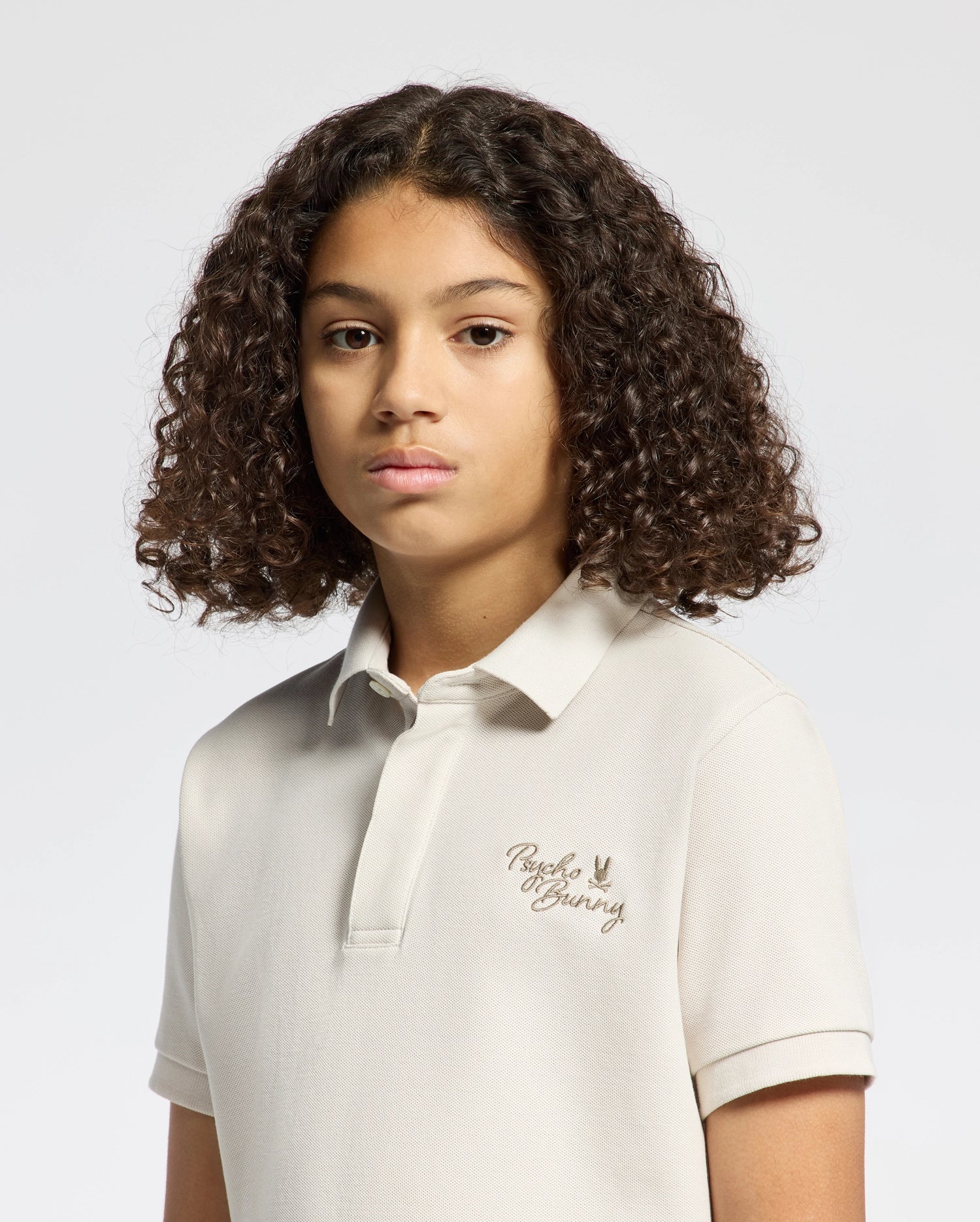 A young person with curly hair wears a Psycho Bunny KIDS DESERT PIQUE POLO - B0K506F200, crafted from luxurious Pima cotton with chest embroidery. They pose against a plain backdrop, looking forward with a neutral expression that captures timeless style.