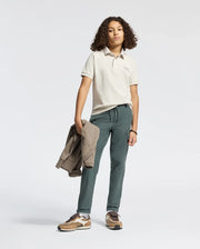 A confident young person with curly hair wears a Psycho Bunny KIDS DESERT PIQUE POLO (B0K506F200) and dark green pants. They hold a jacket, wearing brown sneakers, showcasing timeless style against a plain white studio backdrop.
