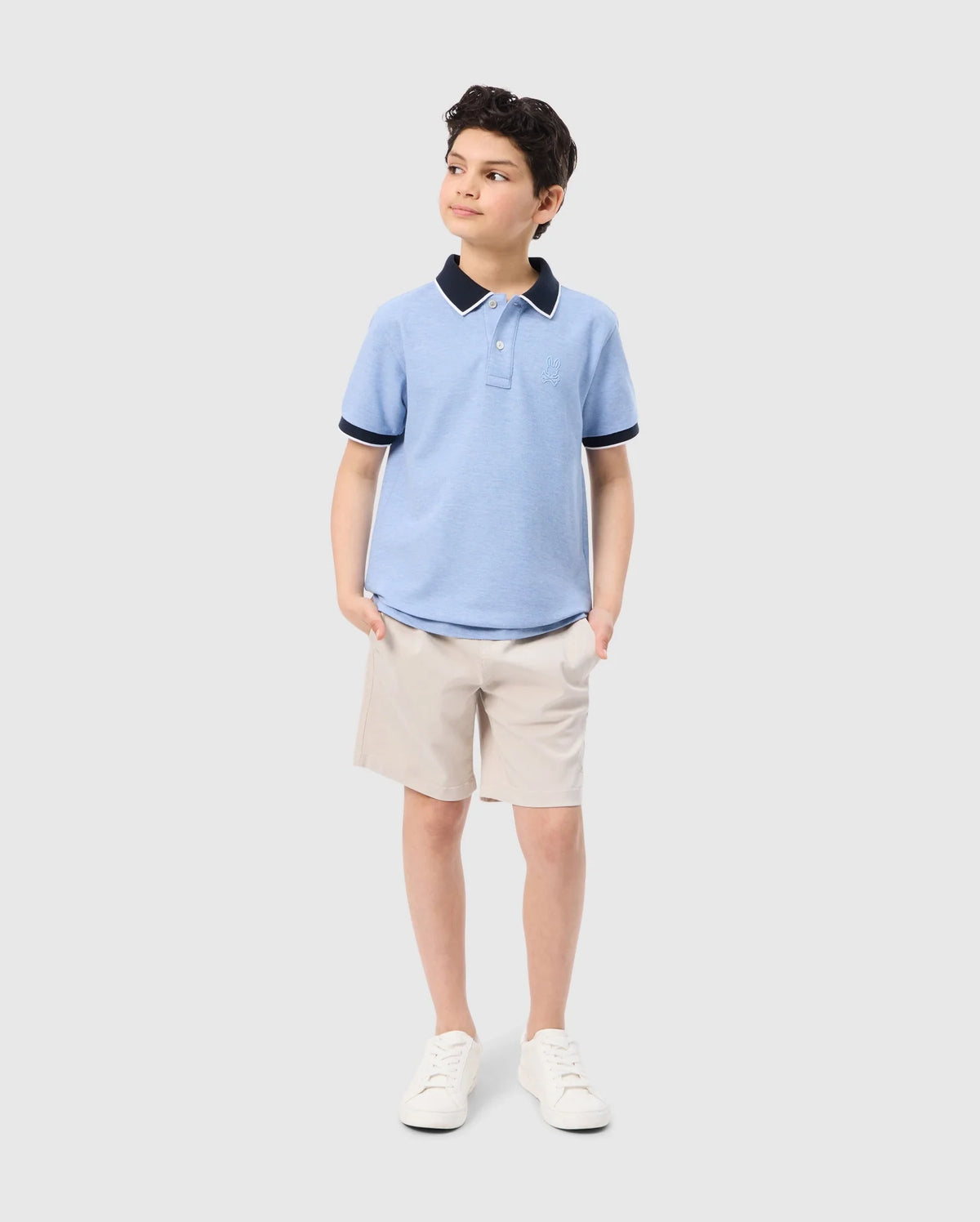 A young boy stands with his hands in his pockets, looking off to the side. He is wearing a Psycho Bunny KIDS WINDCREST PIQUE POLO SHIRT - B0K592C200 featuring a color-blocked collar and khaki shorts. With a slight smile, he completes his look with white sneakers. The background is plain white.