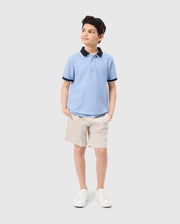 A young boy stands with his hands in his pockets, looking off to the side. He is wearing a Psycho Bunny KIDS WINDCREST PIQUE POLO SHIRT - B0K592C200 featuring a color-blocked collar and khaki shorts. With a slight smile, he completes his look with white sneakers. The background is plain white.