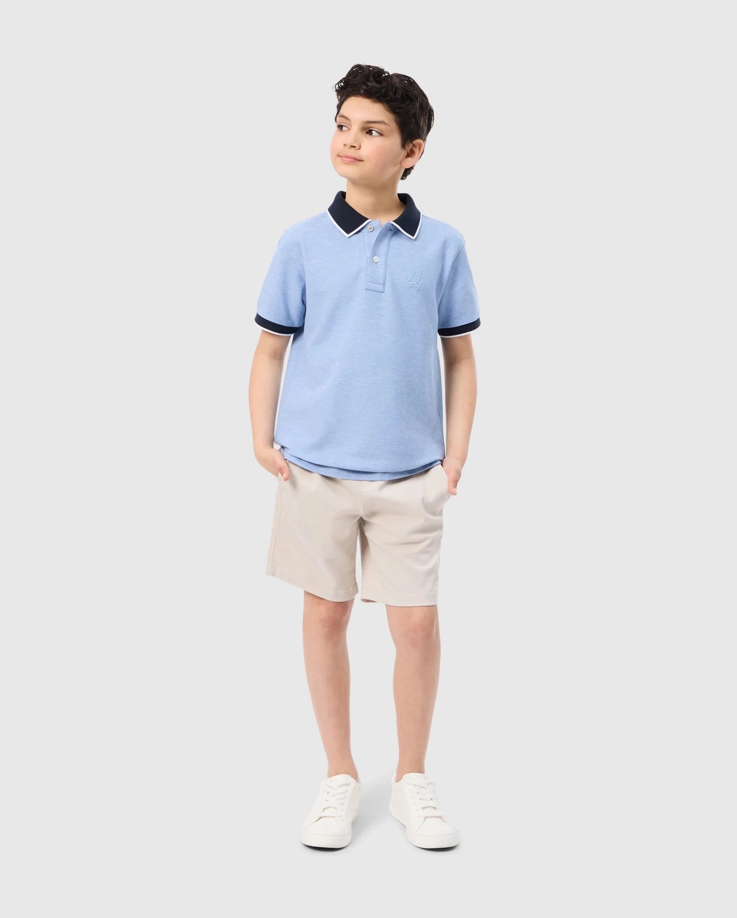 A young boy stands with his hands in his pockets, looking off to the side. He is wearing a Psycho Bunny KIDS WINDCREST PIQUE POLO SHIRT - B0K592C200 featuring a color-blocked collar and khaki shorts. With a slight smile, he completes his look with white sneakers. The background is plain white.