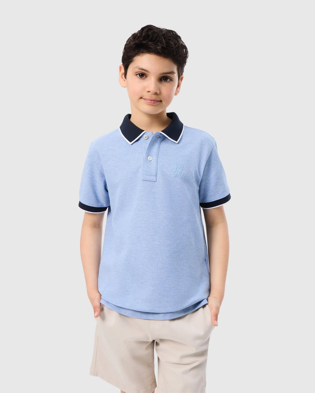 A young boy with curly dark hair stands against a plain white background. He is wearing a light blue KIDS WINDCREST PIQUE POLO SHIRT - B0K592C200 by Psycho Bunny with a striped collar and beige shorts. The boy has one hand in his pocket and a slight smile on his face.
