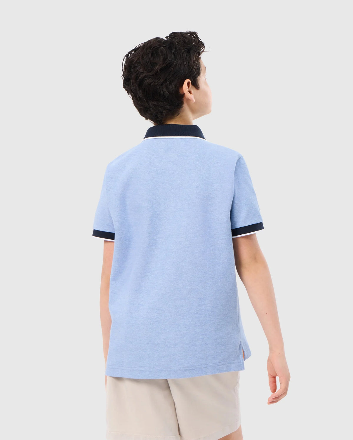 A young person with short, dark hair is seen from the back, wearing a Psycho Bunny KIDS WINDCREST PIQUE POLO SHIRT - B0K592C200 with a color blocked collar and sleeve trim, along with beige shorts. They are standing against a plain white background.