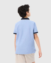 A young person with short, dark hair is seen from the back, wearing a Psycho Bunny KIDS WINDCREST PIQUE POLO SHIRT - B0K592C200 with a color blocked collar and sleeve trim, along with beige shorts. They are standing against a plain white background.