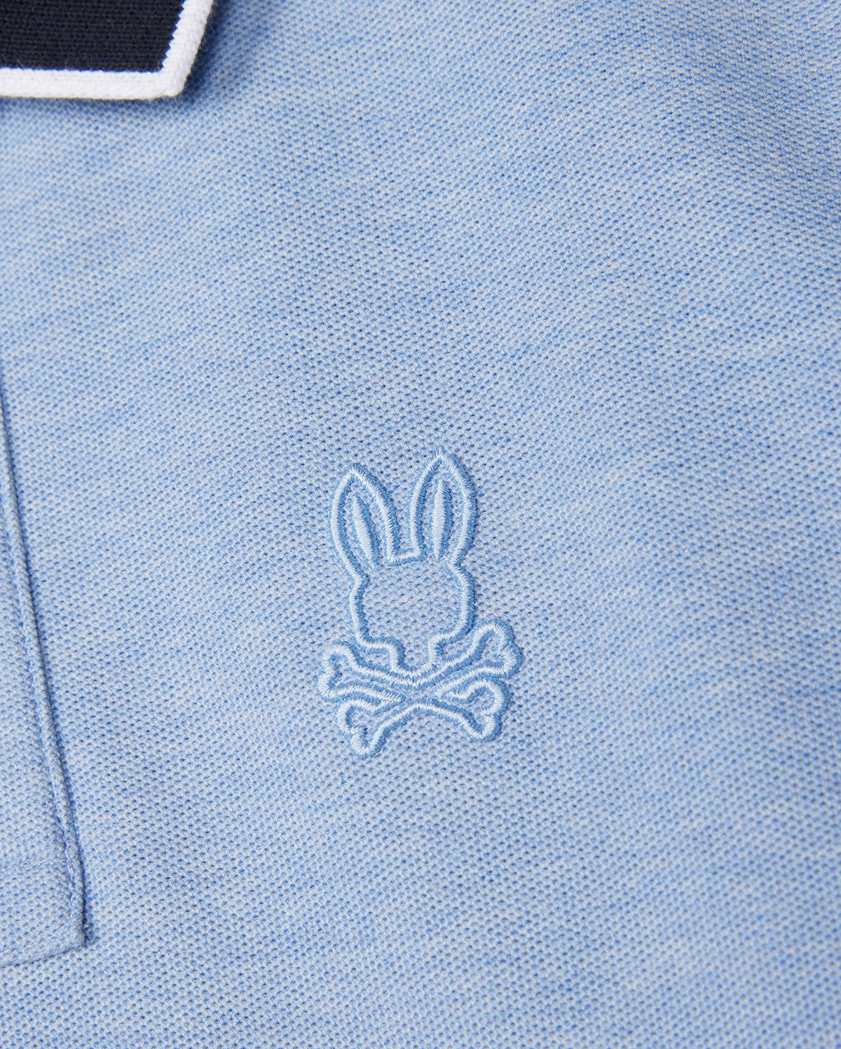 Close-up of a pale blue KIDS WINDCREST PIQUE POLO SHIRT - B0K592C200 from Psycho Bunny with a small, embroidered logo on the chest. The logo features a rabbit's head above two crossed bones, stitched in matching blue thread. The collar has black and white striped edging.