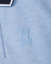 Close-up of a pale blue KIDS WINDCREST PIQUE POLO SHIRT - B0K592C200 from Psycho Bunny with a small, embroidered logo on the chest. The logo features a rabbit's head above two crossed bones, stitched in matching blue thread. The collar has black and white striped edging.