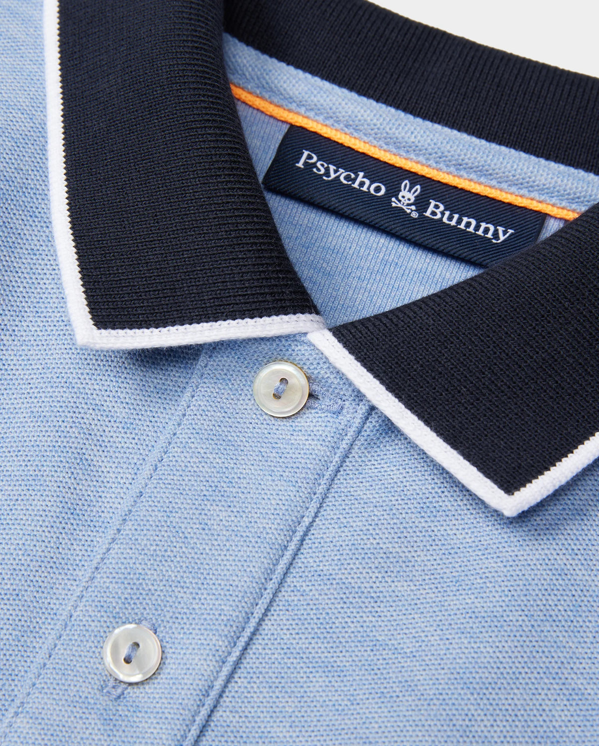Close-up of a KIDS WINDCREST PIQUE POLO SHIRT - B0K592C200 in light blue with a color-blocked black collar featuring white trim. The shirt has two white buttons and a label that reads "Psycho Bunny" with a small raised-embroidered chest Bunny logo.