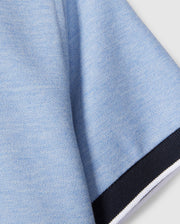 Close-up of a light blue Psycho Bunny KIDS WINDCREST PIQUE POLO SHIRT - B0K592C200 fabric with a textured appearance. The image shows a section of the fabric with a black border along one edge, which includes small white and blue stripes. The background is plain and light-colored.