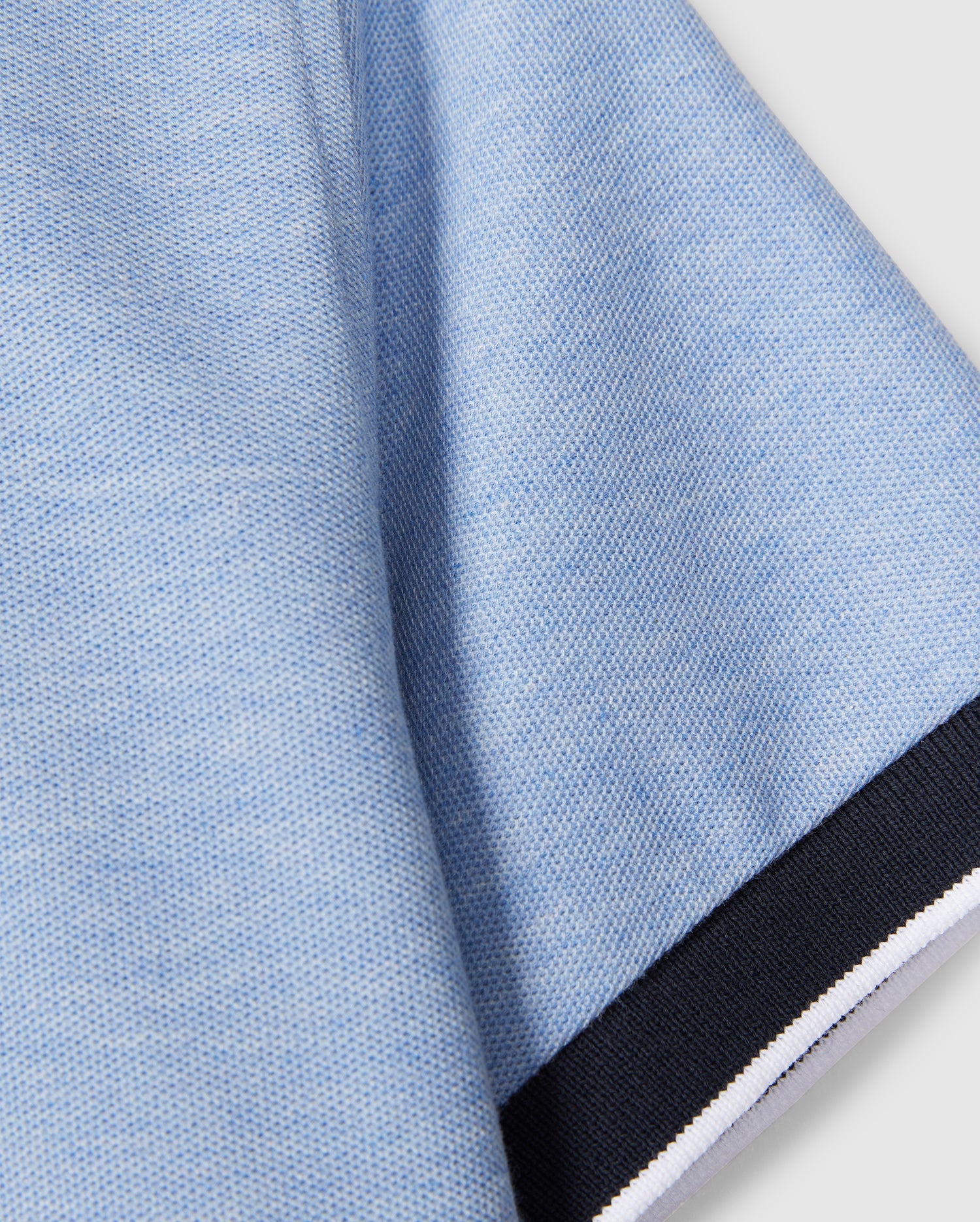 Close-up of a light blue Psycho Bunny KIDS WINDCREST PIQUE POLO SHIRT - B0K592C200 fabric with a textured appearance. The image shows a section of the fabric with a black border along one edge, which includes small white and blue stripes. The background is plain and light-colored.