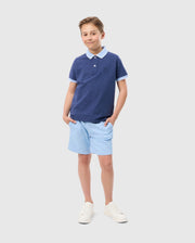 A young boy stands smiling with his hands in his light blue shorts pockets. He is wearing a dark blue Psycho Bunny KIDS WINDCREST PIQUE POLO SHIRT - B0K592C200 with an embroidered chest and a light blue striped collar, paired with white sneakers. The background is plain white.
