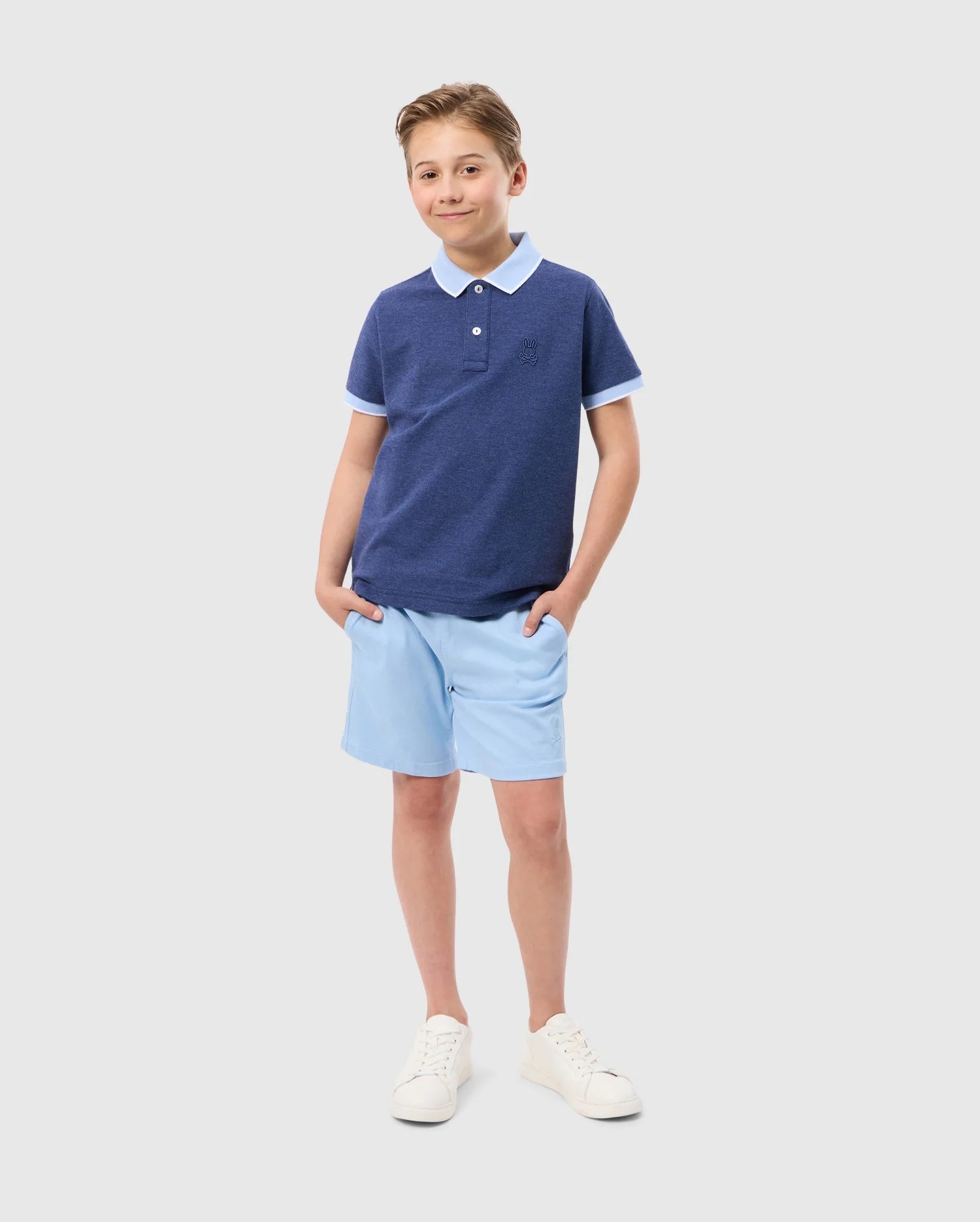 A young boy stands smiling with his hands in his light blue shorts pockets. He is wearing a dark blue Psycho Bunny KIDS WINDCREST PIQUE POLO SHIRT - B0K592C200 with an embroidered chest and a light blue striped collar, paired with white sneakers. The background is plain white.