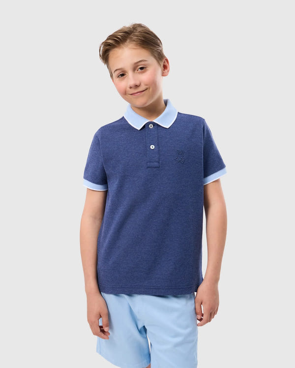 A young boy with light brown hair is smiling, wearing a Psycho Bunny KIDS WINDCREST PIQUE POLO SHIRT - B0K592C200 with a striped collar and light blue sleeve trim. Paired with light blue shorts, he stands against a plain, light-colored background.