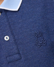 Close-up of a Psycho Bunny KIDS WINDCREST PIQUE POLO SHIRT - B0K592C200 with a light blue striped collar. The shirt has two buttons and features an embroidered logo of a rabbit's head with crossed bones underneath on the left side of the chest.