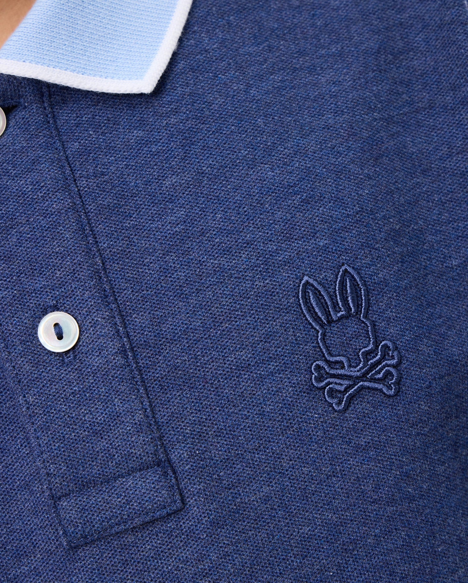 Close-up of a Psycho Bunny KIDS WINDCREST PIQUE POLO SHIRT - B0K592C200 with a light blue striped collar. The shirt has two buttons and features an embroidered logo of a rabbit's head with crossed bones underneath on the left side of the chest.