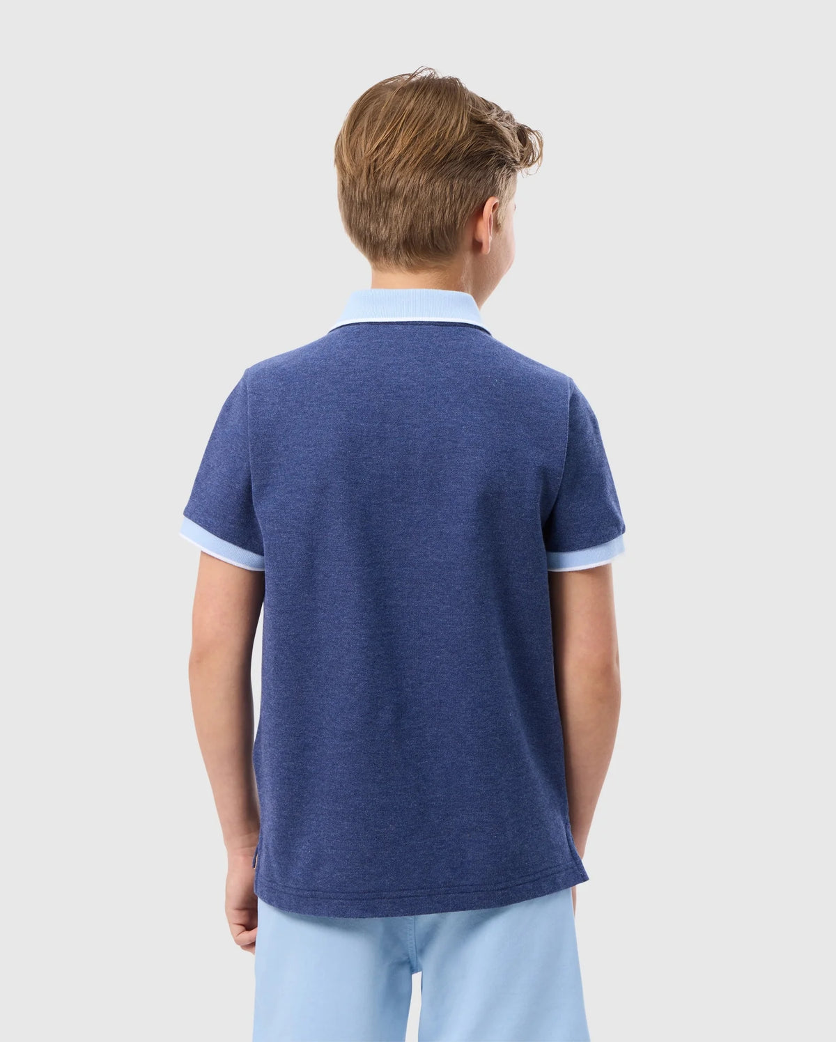 A young boy with light brown hair is standing with his back to the camera. He is wearing a blue KIDS WINDCREST PIQUE POLO SHIRT - B0K592C200 by Psycho Bunny with white trim on the sleeves and striped collar, paired with light blue shorts. The background is plain and white.