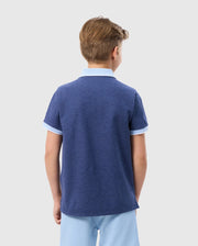A young boy with light brown hair is standing with his back to the camera. He is wearing a blue KIDS WINDCREST PIQUE POLO SHIRT - B0K592C200 by Psycho Bunny with white trim on the sleeves and striped collar, paired with light blue shorts. The background is plain and white.