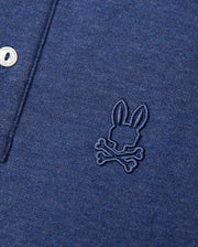 Close-up of a Psycho Bunny KIDS WINDCREST PIQUE POLO SHIRT - B0K592C200 featuring an embroidered rabbit skull and crossbones logo on the left chest. The shirt also has a striped collar and two white buttons partially visible on the left side of the image.