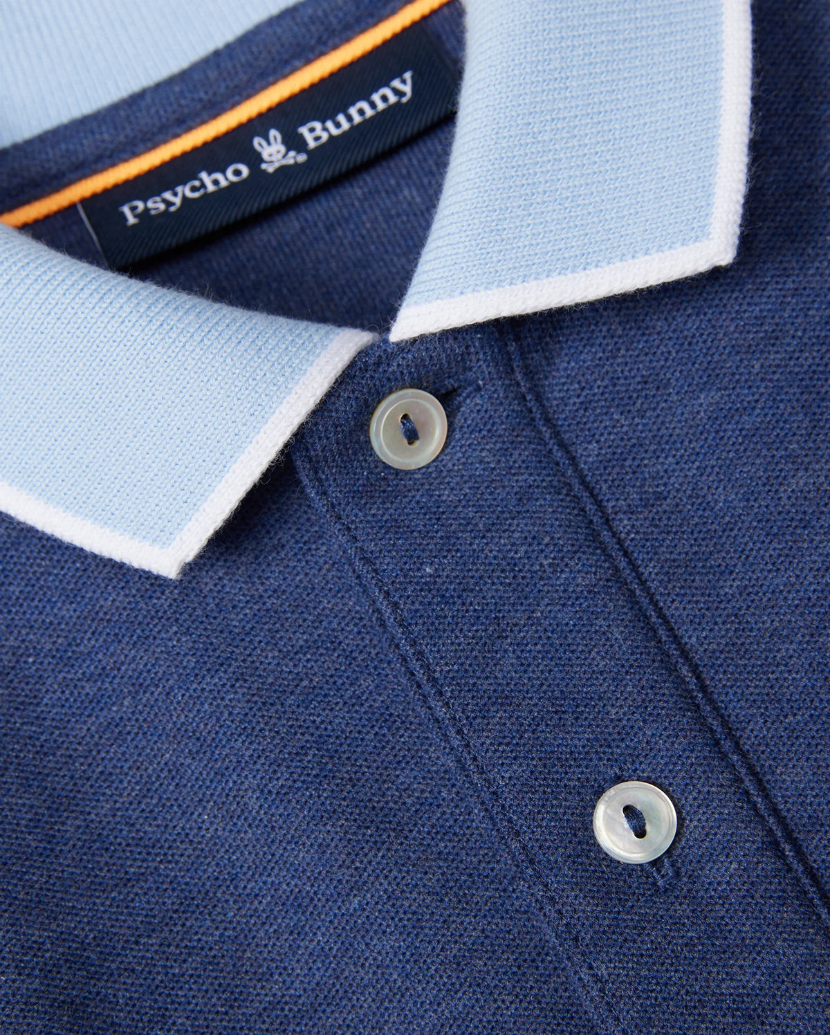 Close-up image of a blue KIDS WINDCREST PIQUE POLO SHIRT - B0K592C200 from Psycho Bunny. The shirt features a light blue striped collar with white trim and two white buttons. The Psycho Bunny label is visible at the back of the collar, along with a small embroidered chest logo of a white bunny.