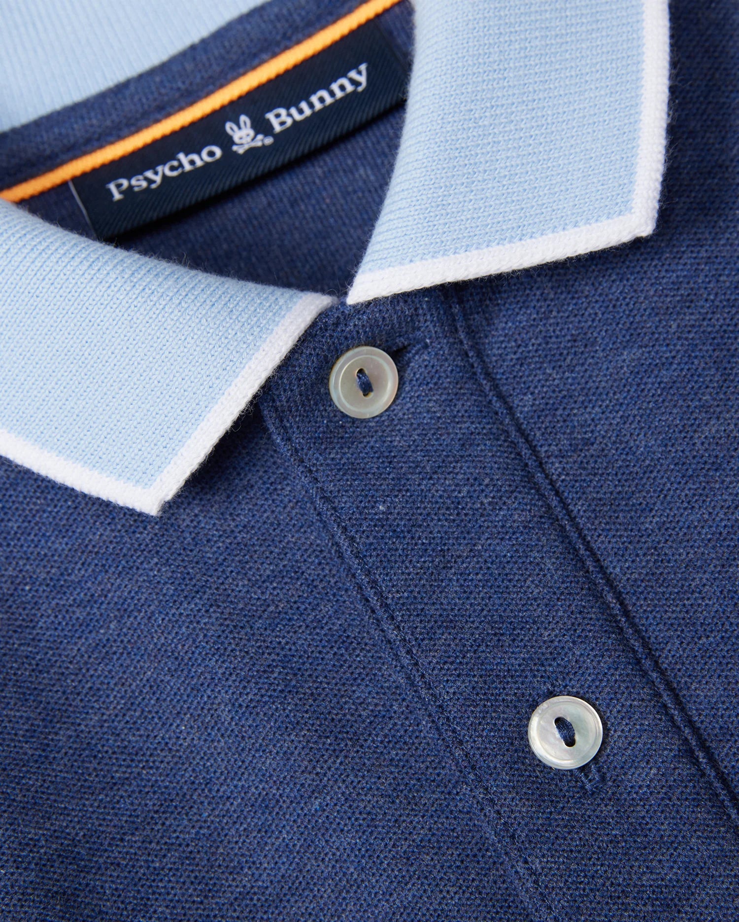 Close-up image of a blue KIDS WINDCREST PIQUE POLO SHIRT - B0K592C200 from Psycho Bunny. The shirt features a light blue striped collar with white trim and two white buttons. The Psycho Bunny label is visible at the back of the collar, along with a small embroidered chest logo of a white bunny.