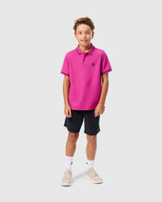 A young boy is smiling at the camera, sporting a bright pink KIDS RIVIERA PIQUE POLO SHIRT - B0K640C200 by Psycho Bunny, paired with dark shorts, white socks, and light-colored sneakers. He stands against a plain background with short, curly brown hair.