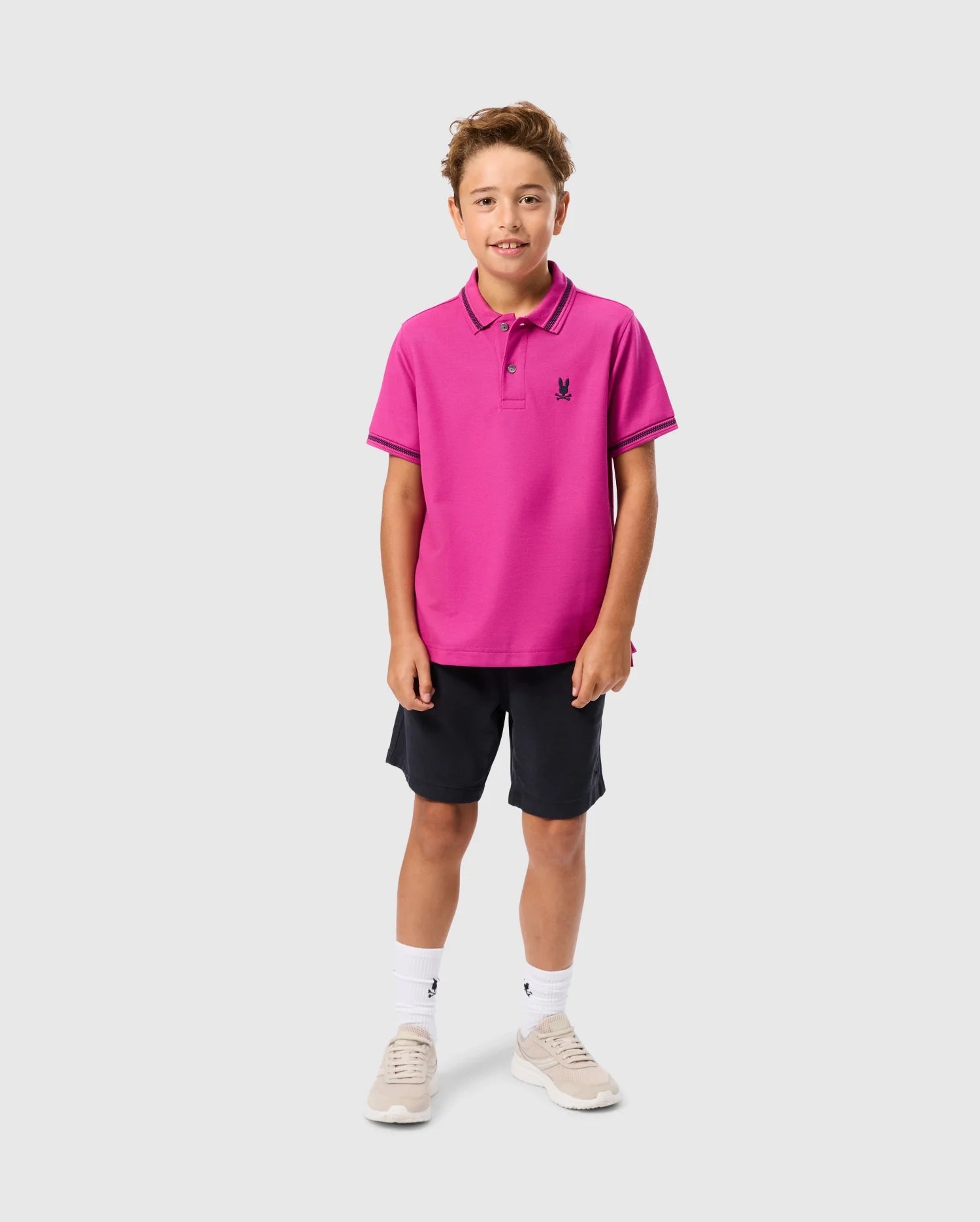 A young boy is smiling at the camera, sporting a bright pink KIDS RIVIERA PIQUE POLO SHIRT - B0K640C200 by Psycho Bunny, paired with dark shorts, white socks, and light-colored sneakers. He stands against a plain background with short, curly brown hair.