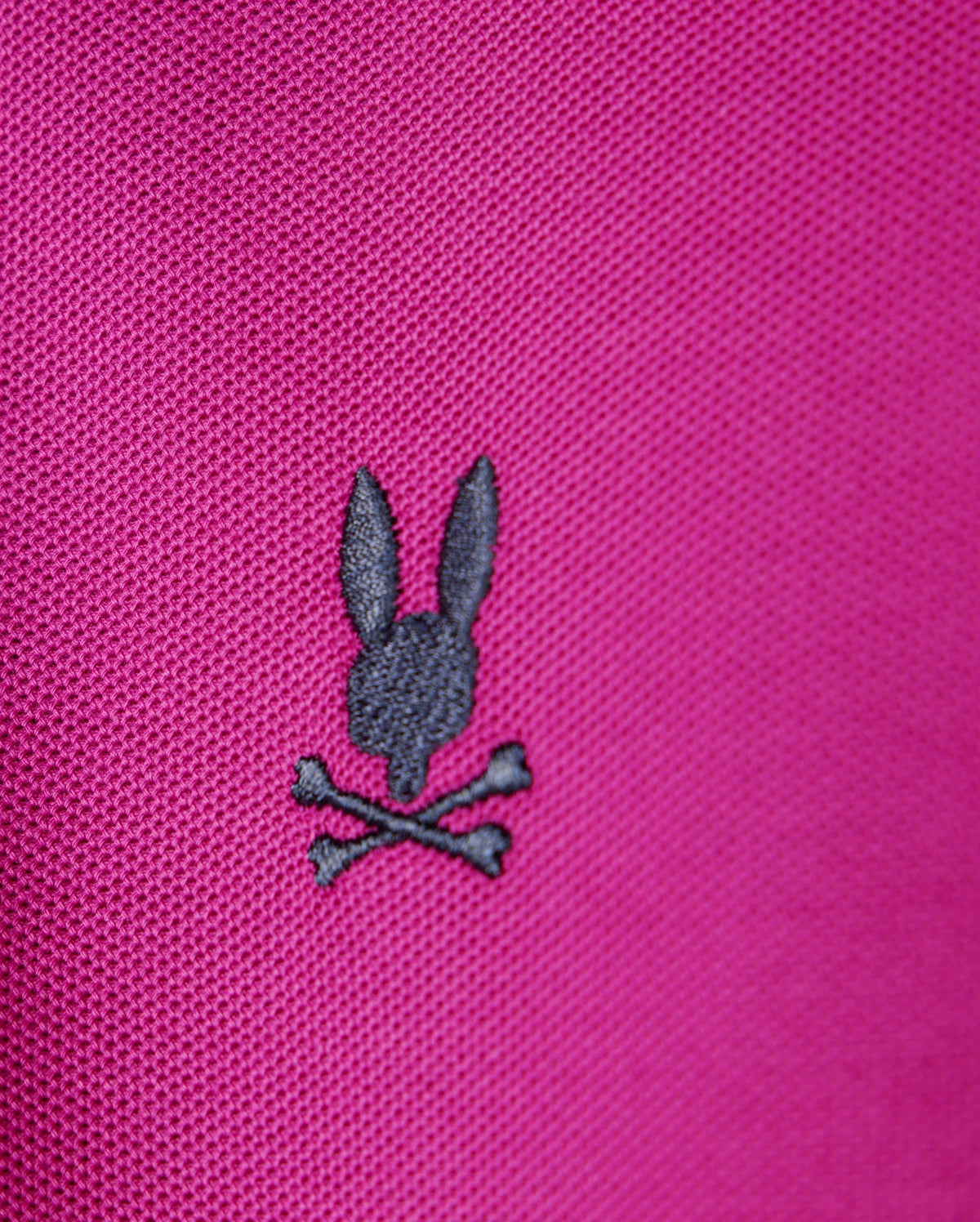 A close-up of the Psycho Bunny KIDS RIVIERA PIQUE POLO SHIRT in pink Pima cotton piqué showcases a small, black embroidered design on the chest. It features a rabbit's head above crossed bones, creating an edgy skull and crossbones motif.