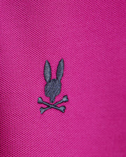 A close-up of the Psycho Bunny KIDS RIVIERA PIQUE POLO SHIRT in pink Pima cotton piqué showcases a small, black embroidered design on the chest. It features a rabbit's head above crossed bones, creating an edgy skull and crossbones motif.