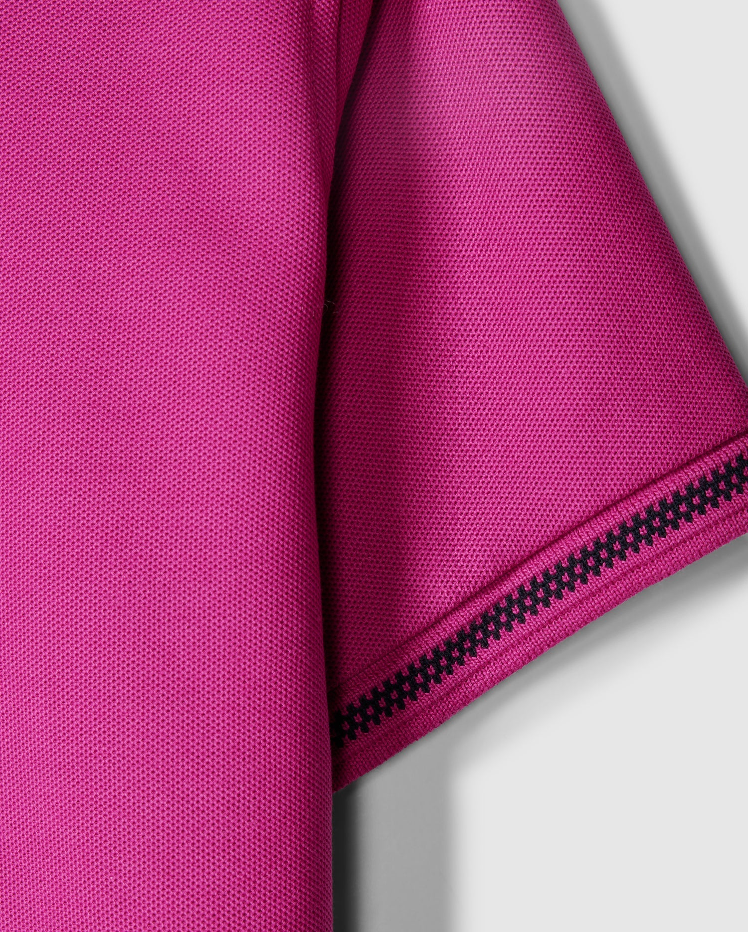 Close-up of a magenta short-sleeve Kids Riviera Pique Polo Shirt by Psycho Bunny, crafted from Pima cotton piqué with a textured fabric. The sleeve is adorned with a black, patterned trim. The background is a plain, light gray color.
