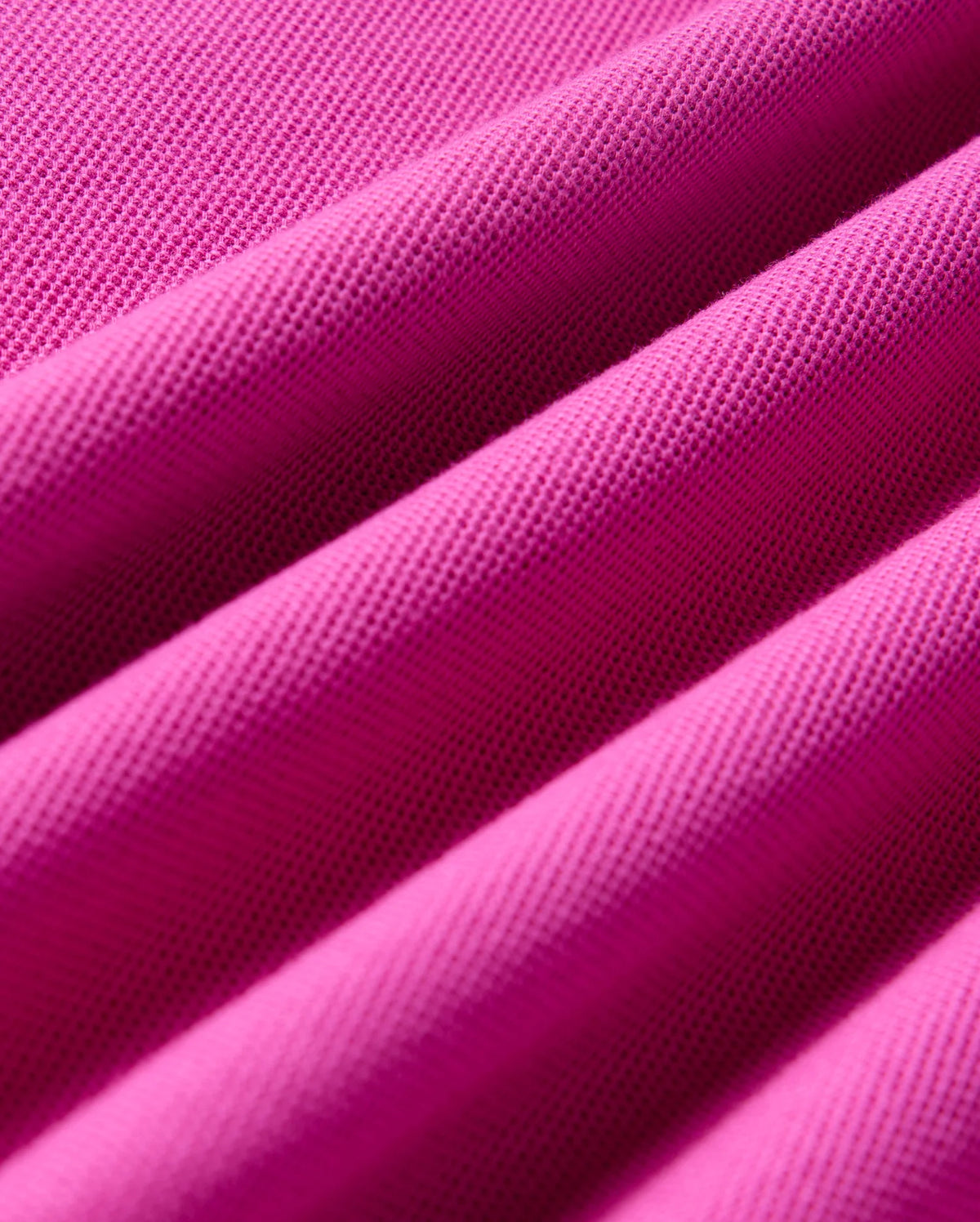 Close-up view of the vibrant pink Pima cotton piqué fabric, featuring a textured, woven pattern from the KIDS RIVIERA PIQUE POLO SHIRT by Psycho Bunny. The fabric is folded into several soft ridges, emphasizing its smooth and structured appearance.