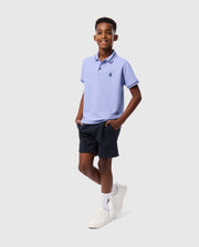 A young boy wearing a Psycho Bunny KIDS RIVIERA PIQUE POLO SHIRT - B0K640C200 made from Pima cotton piqué and navy shorts stands against a light gray background. With his hands in his pockets, he smiles warmly. His outfit is completed with white socks and sneakers, featuring an embroidered chest Bunny for a charming touch.