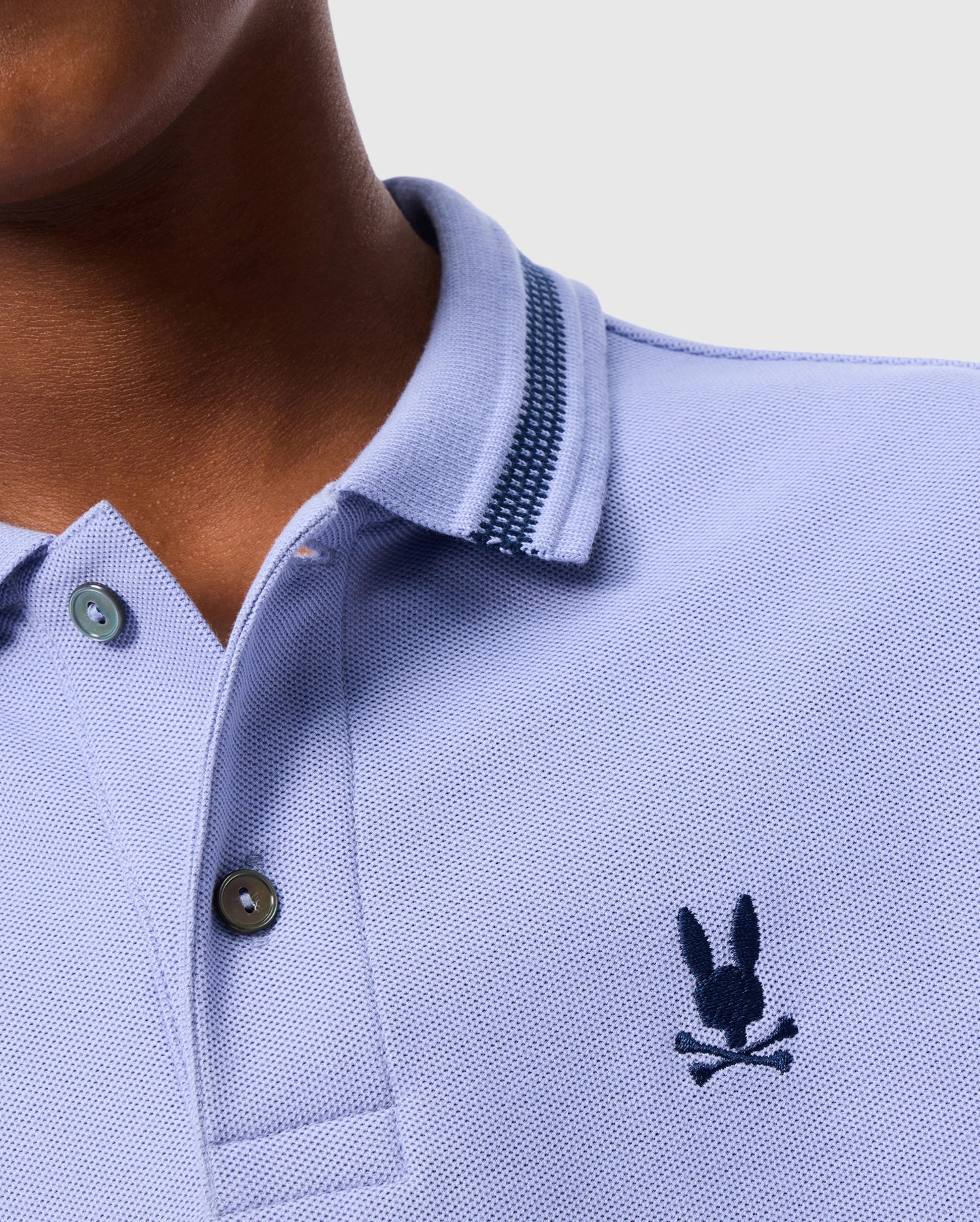 A close-up image shows someone wearing the KIDS RIVIERA PIQUE POLO SHIRT - B0K640C200 from Psycho Bunny in light blue Pima cotton piqué, adorned with an embroidered chest Bunny above crossed bones. The shirt highlights a collar with striped detailing and has two buttons visible.