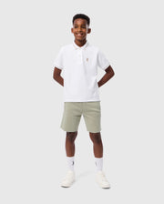 A boy stands against a plain background, wearing a white Psycho Bunny KIDS RIVIERA PIQUE POLO SHIRT - B0K640C200 made of Pima cotton, featuring an embroidered chest Bunny logo. He pairs it with light gray shorts, white socks, and white sneakers. With a smiling expression and his hands behind his back, he looks effortlessly stylish.