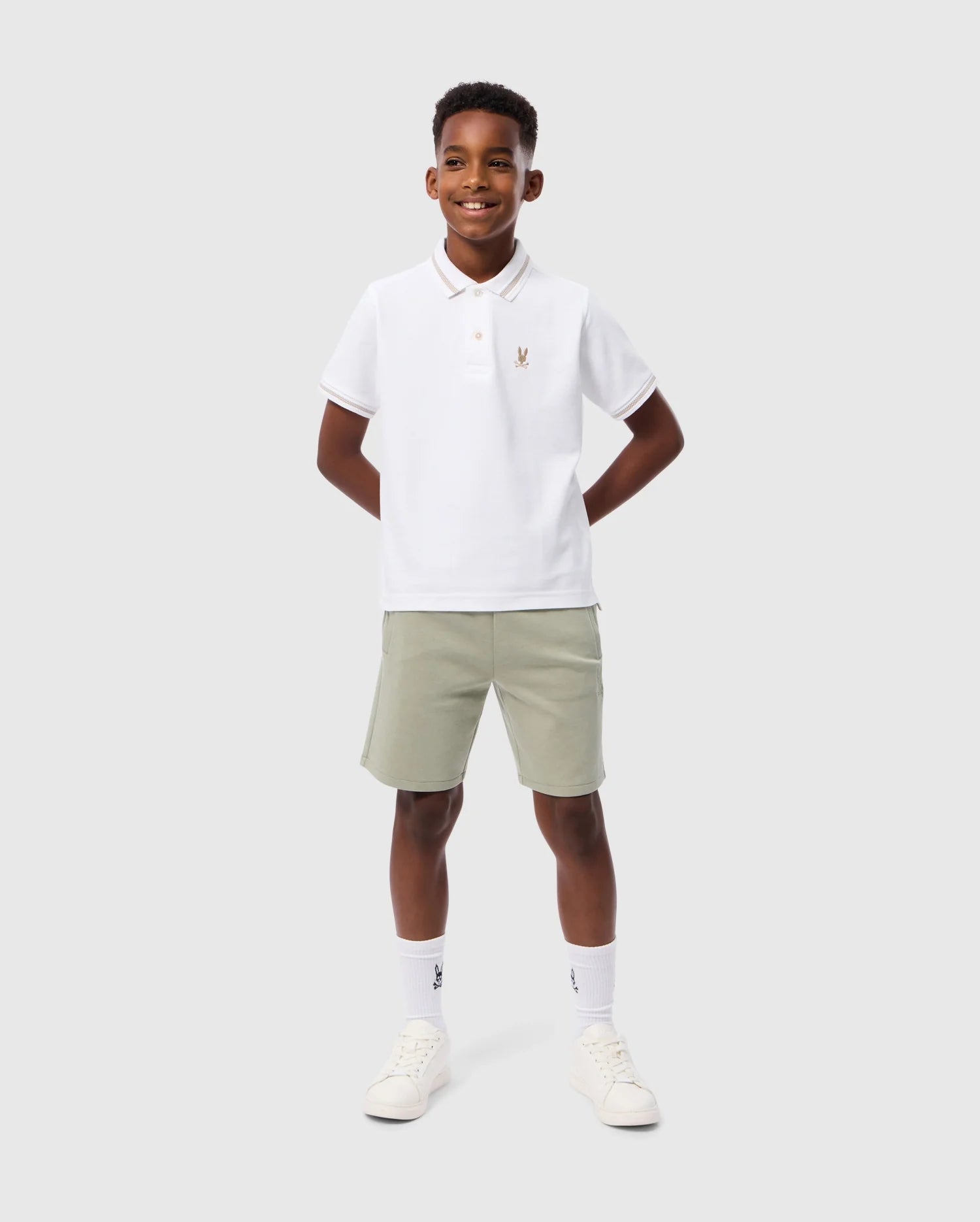 A boy stands against a plain background, wearing a white Psycho Bunny KIDS RIVIERA PIQUE POLO SHIRT - B0K640C200 made of Pima cotton, featuring an embroidered chest Bunny logo. He pairs it with light gray shorts, white socks, and white sneakers. With a smiling expression and his hands behind his back, he looks effortlessly stylish.