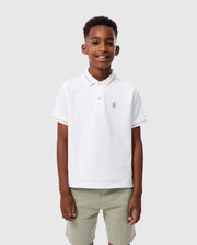 A young boy stands smiling against a light gray backdrop. He is wearing a Psycho Bunny Kids Riviera Pique Polo Shirt - B0K640C200, made from soft Pima cotton, with an embroidered chest Bunny logo on the left, and light green shorts. His hands rest by his sides.