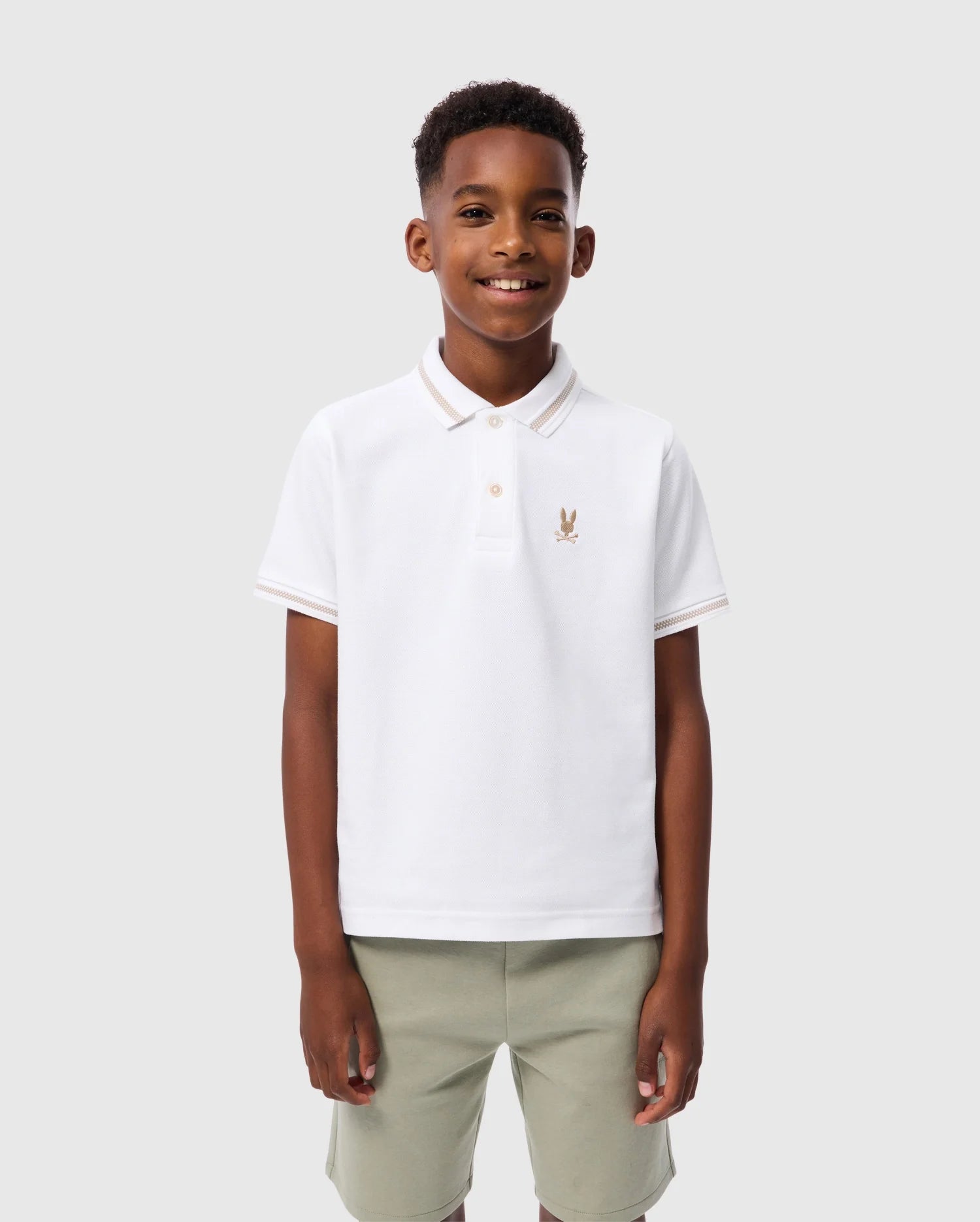 A young boy stands smiling against a light gray backdrop. He is wearing a Psycho Bunny Kids Riviera Pique Polo Shirt - B0K640C200, made from soft Pima cotton, with an embroidered chest Bunny logo on the left, and light green shorts. His hands rest by his sides.