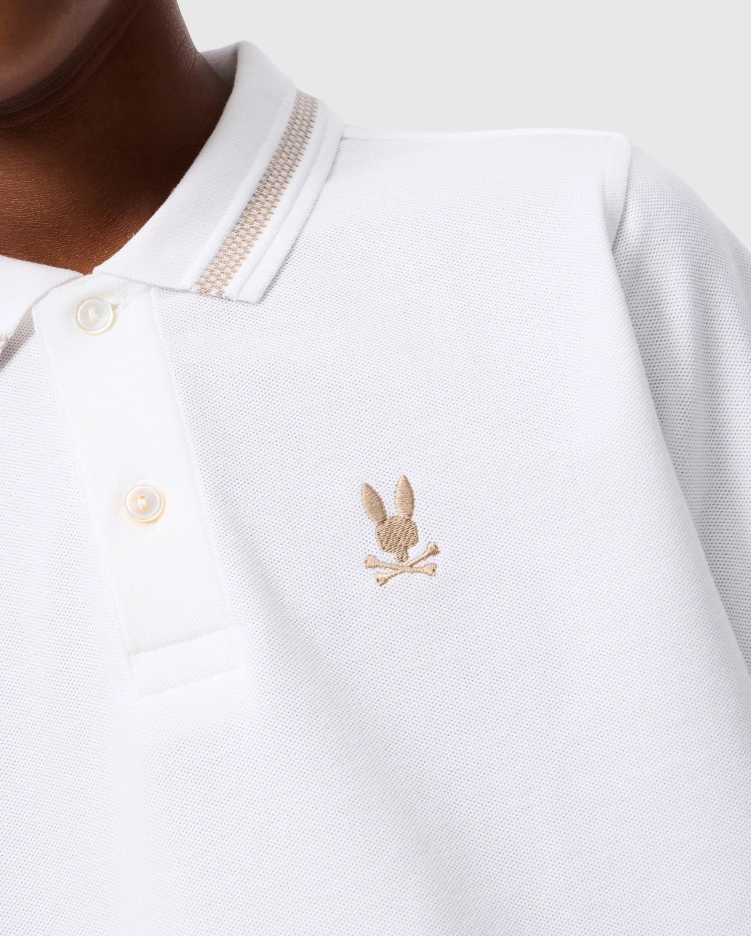 Here is a detailed close-up of a person wearing the KIDS RIVIERA PIQUE POLO SHIRT - B0K640C200 by Psycho Bunny in white Pima cotton. The polo features a beige embroidered Bunny head above crossbones on the left chest. It also has a collared neckline with buttons and a striped detail along the collar edge.