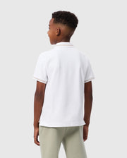 A young boy with short curly hair is standing and facing away from the camera. He is wearing a white short-sleeved KIDS RIVIERA PIQUE POLO SHIRT - B0K640C200 by Psycho Bunny, made of Pima cotton, paired with light gray pants. The background is plain and light gray.